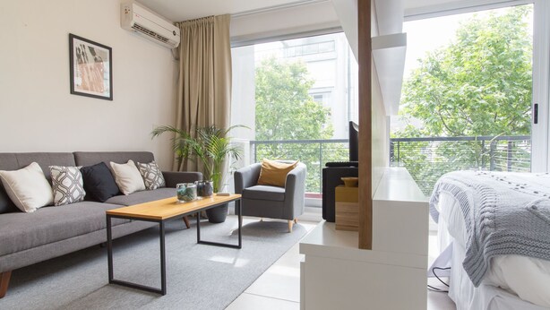 Een moderne woonkamer met een grijze bank, een houten salontafel, een grijze fauteuil en een wit bed. Er is een groot raam met uitzicht op bomen en een balkon.