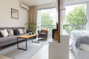 Un salon moderne avec un canapé gris, une table basse en bois, un fauteuil gris et un lit blanc. Une grande fenêtre offre une vue sur les arbres, et il y a un balcon.
