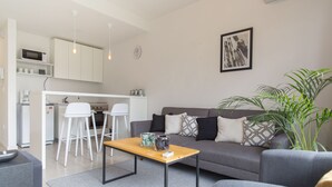 Een moderne woonkamer met een grijze bank, een houten salontafel en een grijze fauteuil. Er is een keuken met witte kastjes en een magnetron.