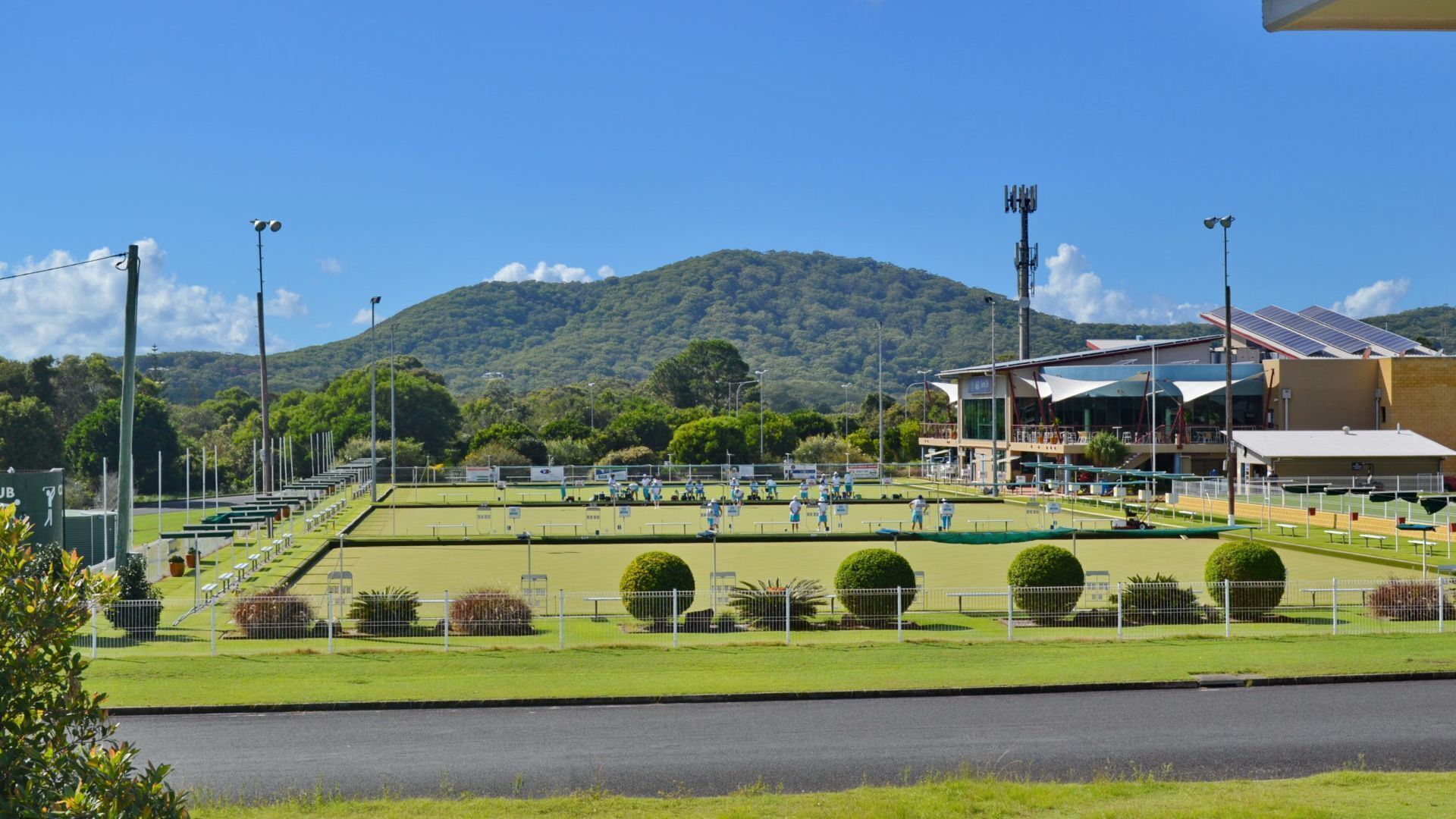 A tennis court with players, surrounded by a fence, with a building and hills in the background.