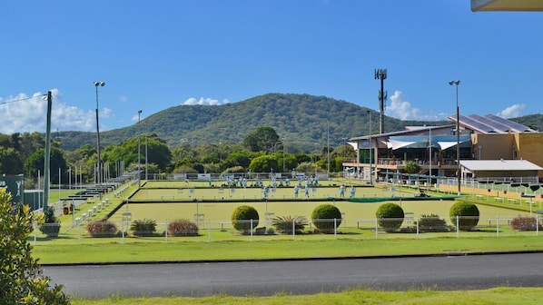 A tennis court with players, surrounded by a fence, with a building and hills in the background.