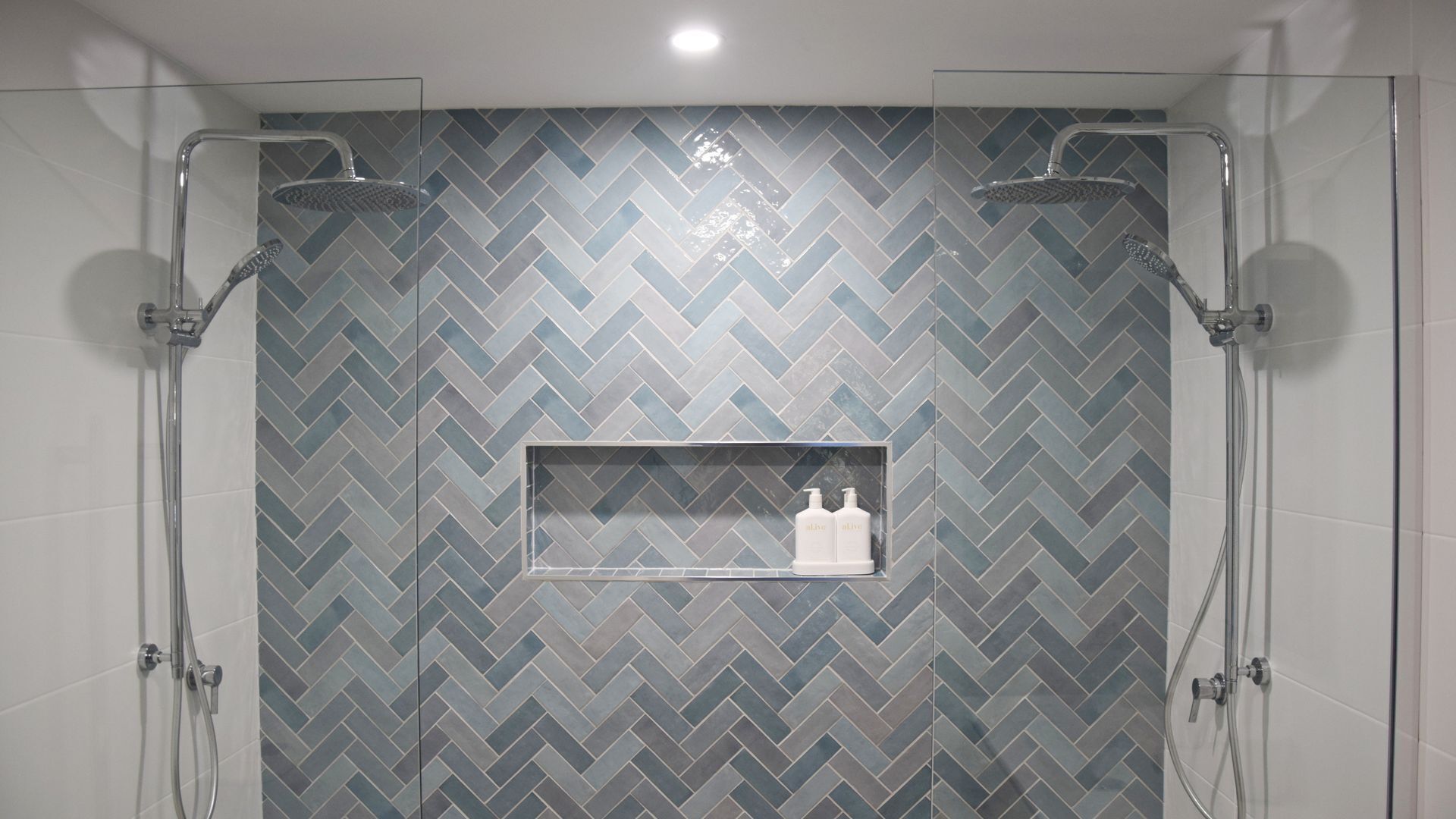 Un espace douche avec un carrelage disposé en motif chevron, une étagère en verre pour les articles de toilette et deux pommes de douche au plafond.