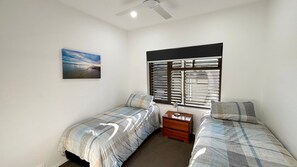 A bedroom with two beds, a nightstand, a ceiling fan, a window with blinds, and a painting on the wall. - Waterford Apartment 3 - Bring your own linen (South West Rocks)