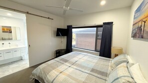 A bedroom with a bed, a window with curtains, a flat-screen TV, a bedside table, and a bathroom with a mirror and sink.