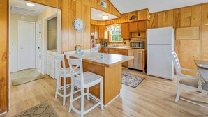 A compact kitchen with wooden walls, a central island with chairs, a refrigerator, a microwave, and a clock on the wall.