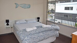 A bedroom with a bed, two bedside lamps, a window with a view of buildings, and a wall decoration of a fish.