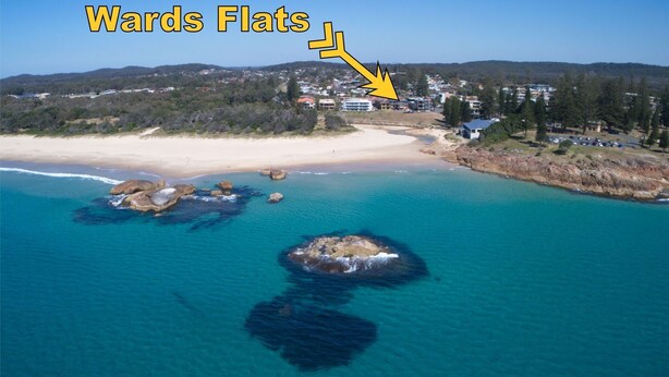 An aerial view of a coastal area with a beach, a rocky outcrop in the water, and a residential area with houses and a road.