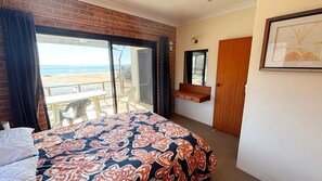 A bedroom with a large bed, a wooden bench, a door, and a view of the beach. - Main Beach 1 - beachfront (South West Rocks)
