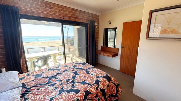 A bedroom with a large bed, a wooden bench, a door, and a view of the beach. - Main Beach 1 - beachfront (South West Rocks)