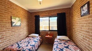 A small room with two single beds, a window with curtains, a wall-mounted picture, and a brick wall. - Main Beach 1 - beachfront - Bring your own linen (South West Rocks)