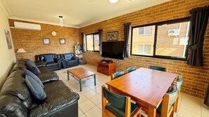 A living room with a brick wall, a sofa, a TV, a table, and chairs. - Main Beach 1 - beachfront (South West Rocks)