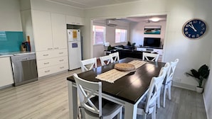 A modern kitchen with white cabinetry, a built-in dishwasher, and a dining area with a dark wooden table and white chairs. - Amble Inn - walk to the beach (SOUTH WEST ROCKS)