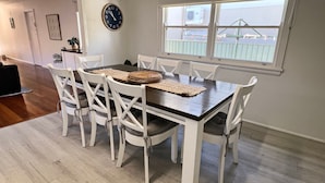 A dining area with a wooden table and white chairs, a clock on the wall, and a window with a view of a fence. - Amble Inn - walk to the beach (SOUTH WEST ROCKS)