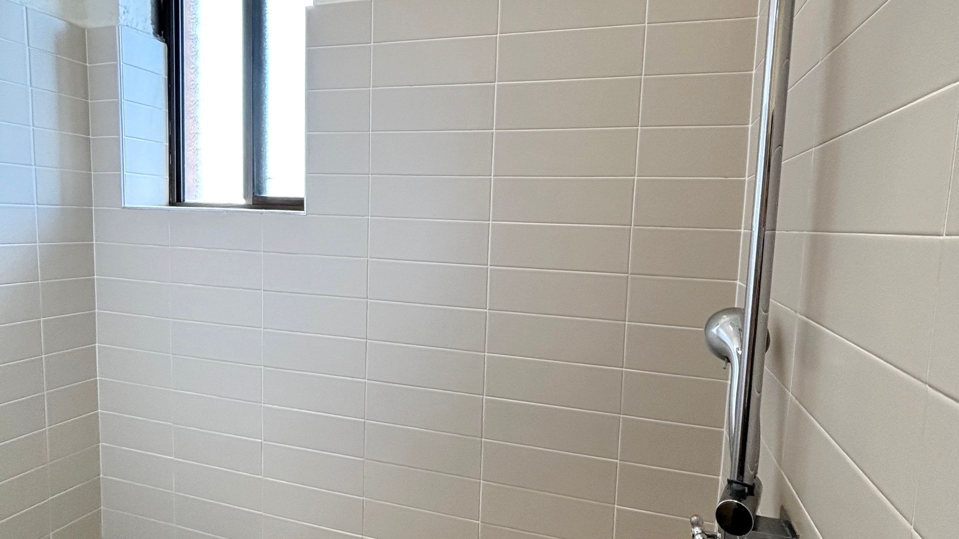 A bathroom with white tiled walls, a window, and a shower with a chrome handle.