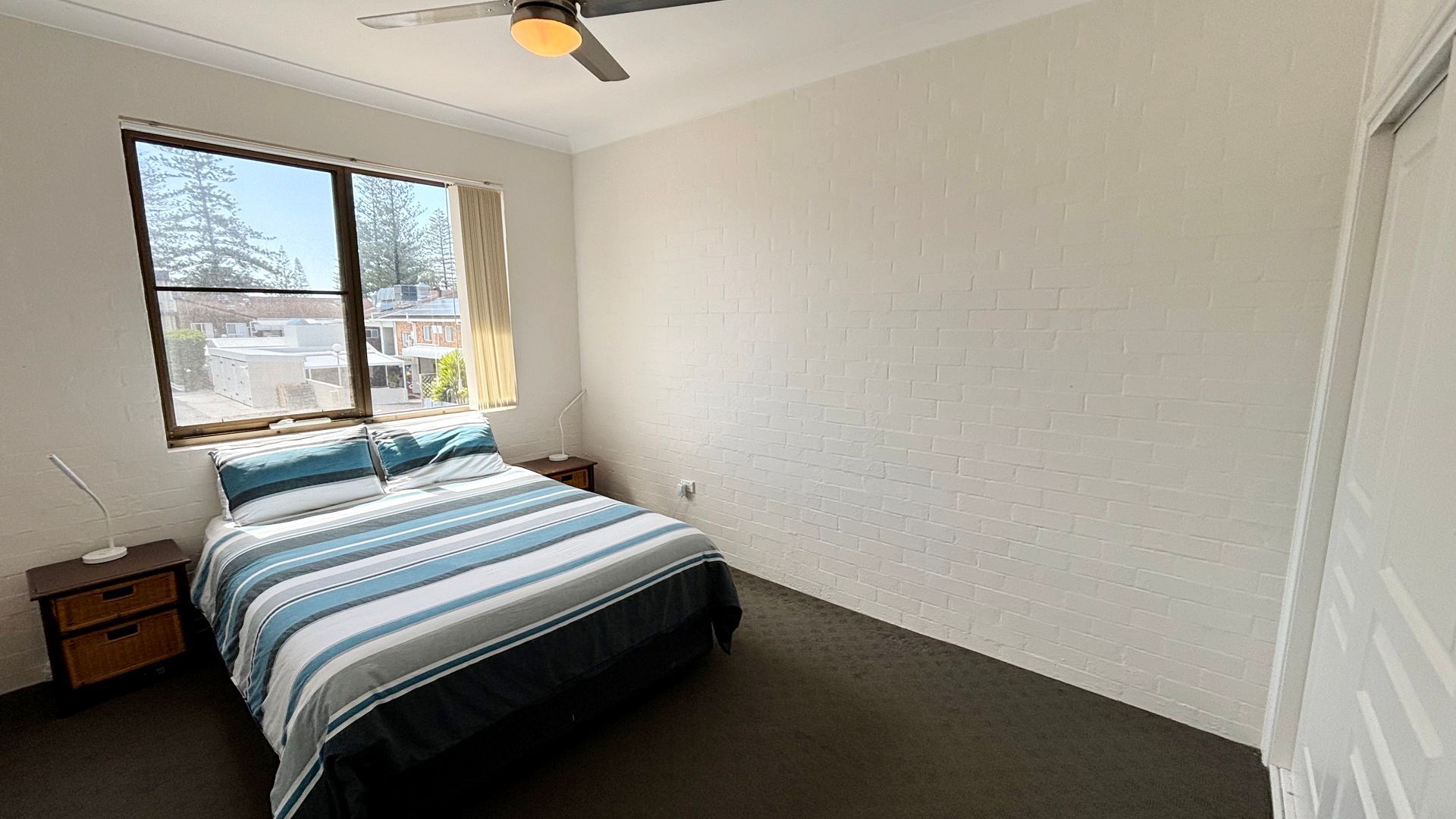 Une chambre à coucher avec un lit, une table de chevet, une lampe, une fenêtre et un ventilateur de plafond.