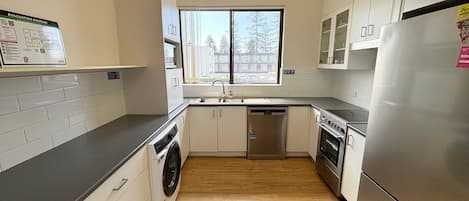 A compact kitchen with a washing machine, a dishwasher, a refrigerator, and a window with a view of trees.