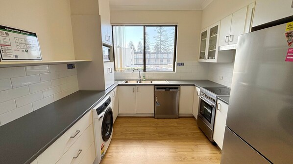 A compact kitchen with a washing machine, a dishwasher, a refrigerator, and a window with a view of trees.
