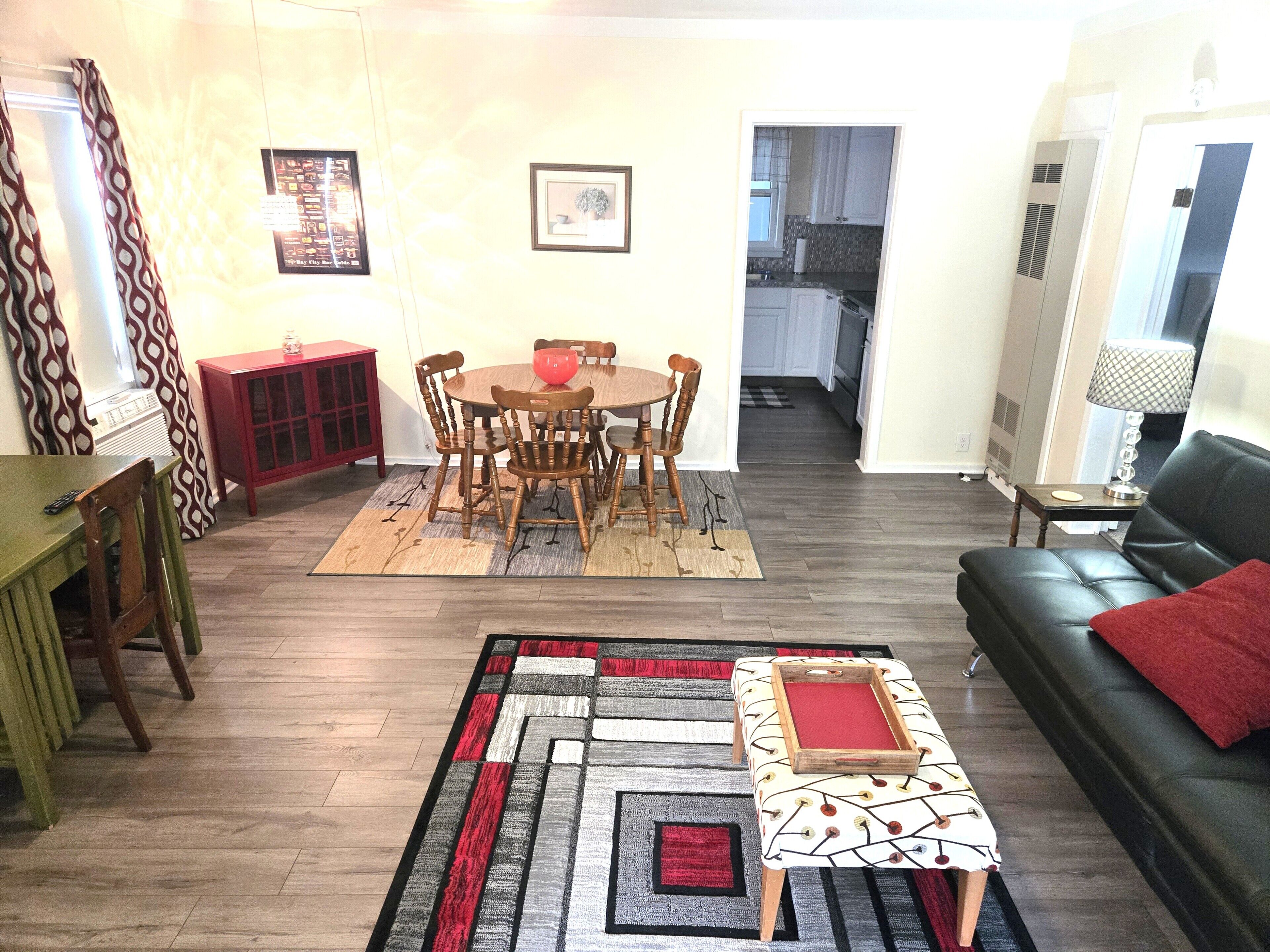 A living room with a sofa, a dining table with chairs, a red cabinet, a patterned rug, and a small ottoman.