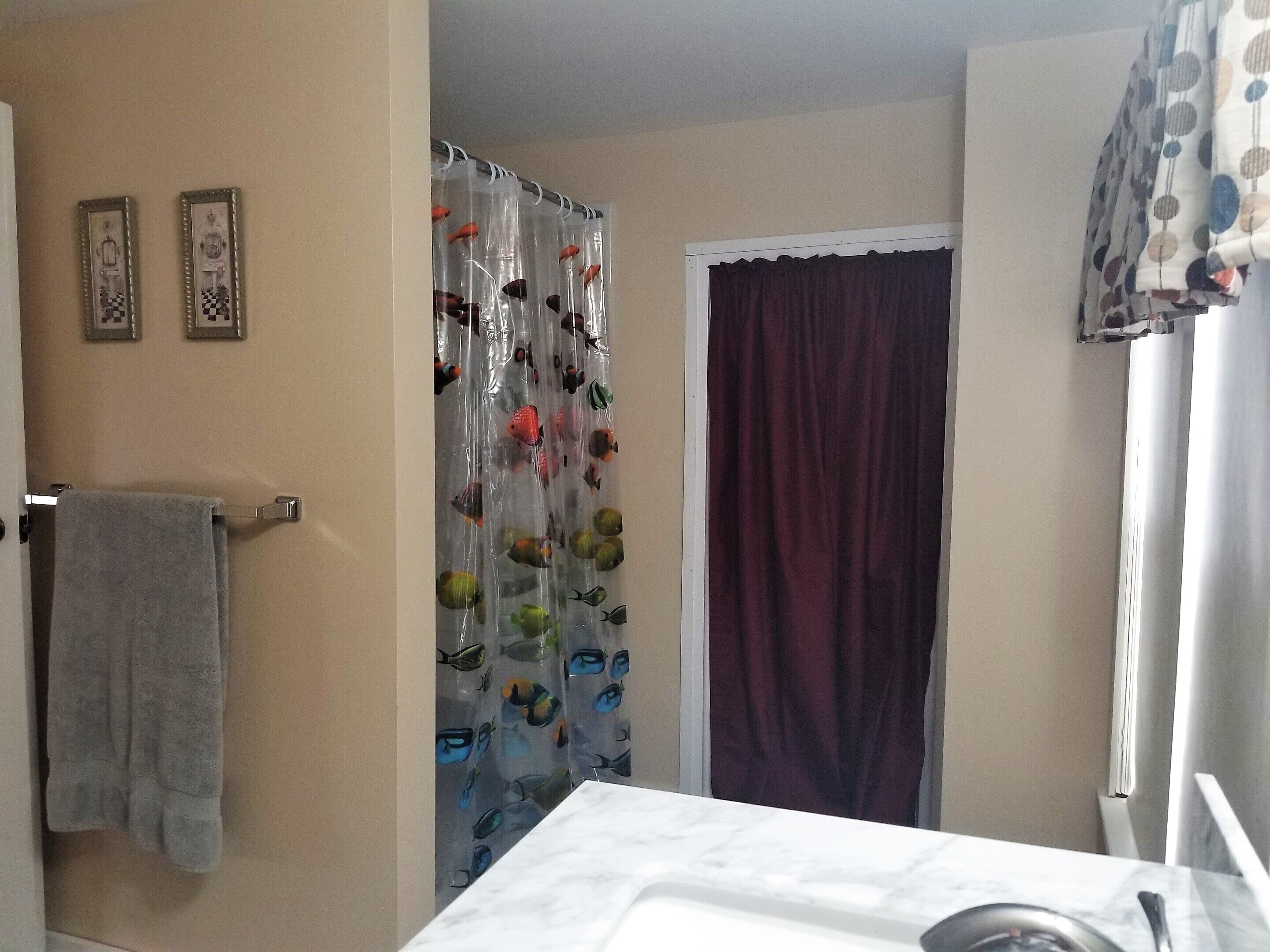 Une salle de bain avec un rideau de douche orné de motifs de poissons, un porte-serviettes avec une serviette grise et une fenêtre avec un rideau.