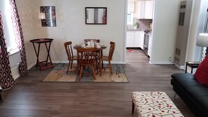 A dining area with a round table and chairs, a small side table, and a bench. A kitchen is visible in the background.