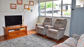 A living room with a television on a wooden stand, two patterned armchairs, a grey rug, and a window with a view of the outside. - Sunshine Cottage - walk to the beach - Bring your own linen (SOUTH WEST ROCKS)