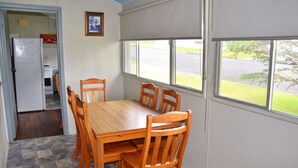 A dining area with a wooden table and chairs, a kitchen with a refrigerator, and a window with a view of the outdoors. - Sunshine Cottage - walk to the beach - Bring your own linen (SOUTH WEST ROCKS)