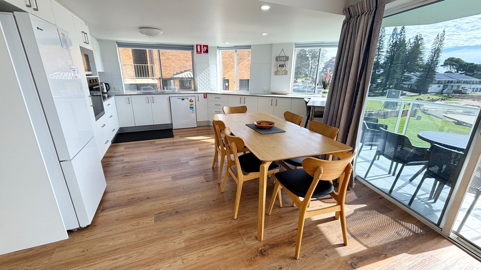 A modern kitchen with white cabinets, a wooden dining table with chairs, and a large window with a view of a grassy area.