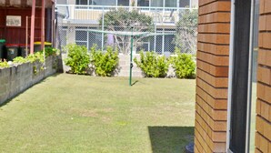 A fenced outdoor area with a grassy lawn, a metal structure, and a brick wall. - Pacific Court 2 - walk to the beach - Linen supplied (SOUTH WEST ROCKS)