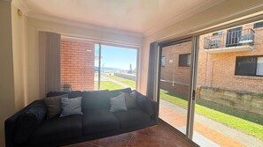 Un balcón con puerta de cristal, un sofá negro con cojines, y vista a un edificio de ladrillo y una zona verde.