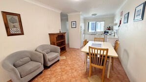 A compact kitchen with a dining table and two chairs, a small wooden cabinet, and a tiled floor. - Pacific Court 2 - walk to the beach - Linen supplied (SOUTH WEST ROCKS)