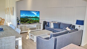 A modern living room with a large flat-screen TV, a sofa set, a coffee table, and a dining area with wicker chairs. - Joanna Cottage - walk to the beach (SOUTH WEST ROCKS)