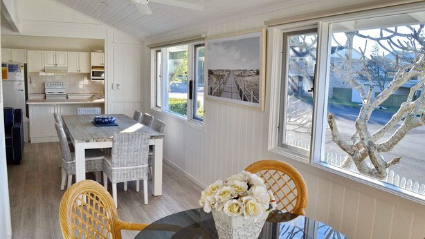 A dining area with a table, chairs, and a vase of flowers. There is a kitchen visible in the background. - Joanna Cottage - walk to the beach (SOUTH WEST ROCKS)