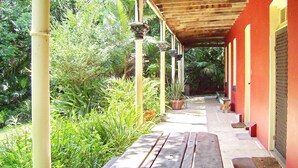 A covered outdoor area with a wooden bench, potted plants, and a building with a red wall and a metal gate.