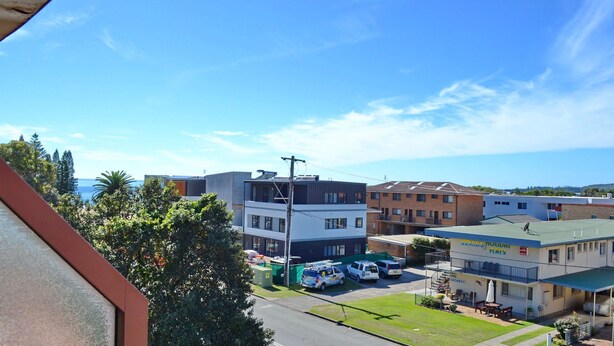Um edifício de vários andares com telhado plano, área de estacionamento com vários veículos e vista para o mar ao fundo.