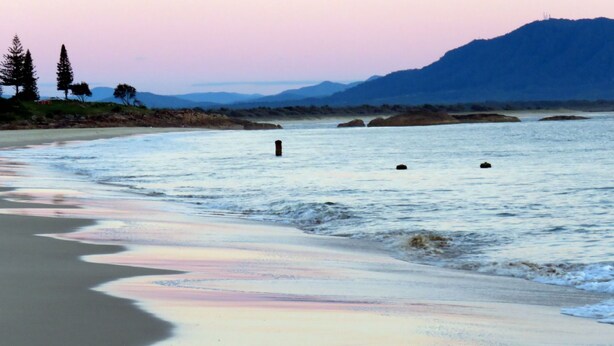 Ein Strand mit ruhigem Wasser, sandigem Ufer und Bergen im Hintergrund.