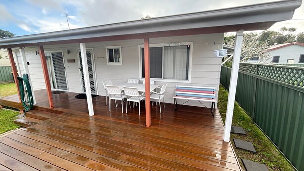 A covered outdoor area with a wooden deck, white plastic chairs, a bench, and a small table. - Wenlaw Cottage (Rear) Hat Head (HAT HEAD)