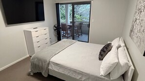 Une chambre à coucher comprenant un lit, une commode, un téléviseur et une porte-fenêtre coulissante donnant sur un balcon équipé de chaises.