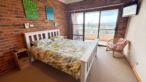 A bedroom with a bed, a small wooden side table, a wicker chair, and a balcony with a view. - Seaview Townhouse 2 - walk to the beach (SOUTH WEST ROCKS)