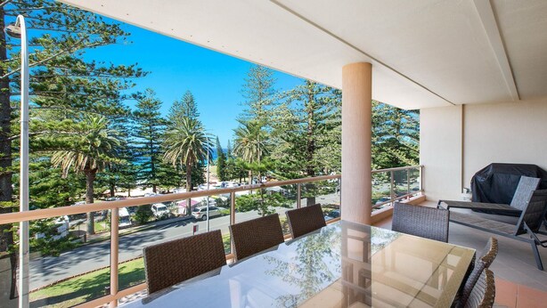A balcony with a glass table and chairs, overlooking a parking area and trees. - Astor Apartment 2 - walk to the beach (SOUTH WEST ROCKS)