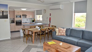 A modern kitchen with a dining area, a gray sofa, and a view of greenery outside. - Astor Apartment 2 - walk to the beach (SOUTH WEST ROCKS)