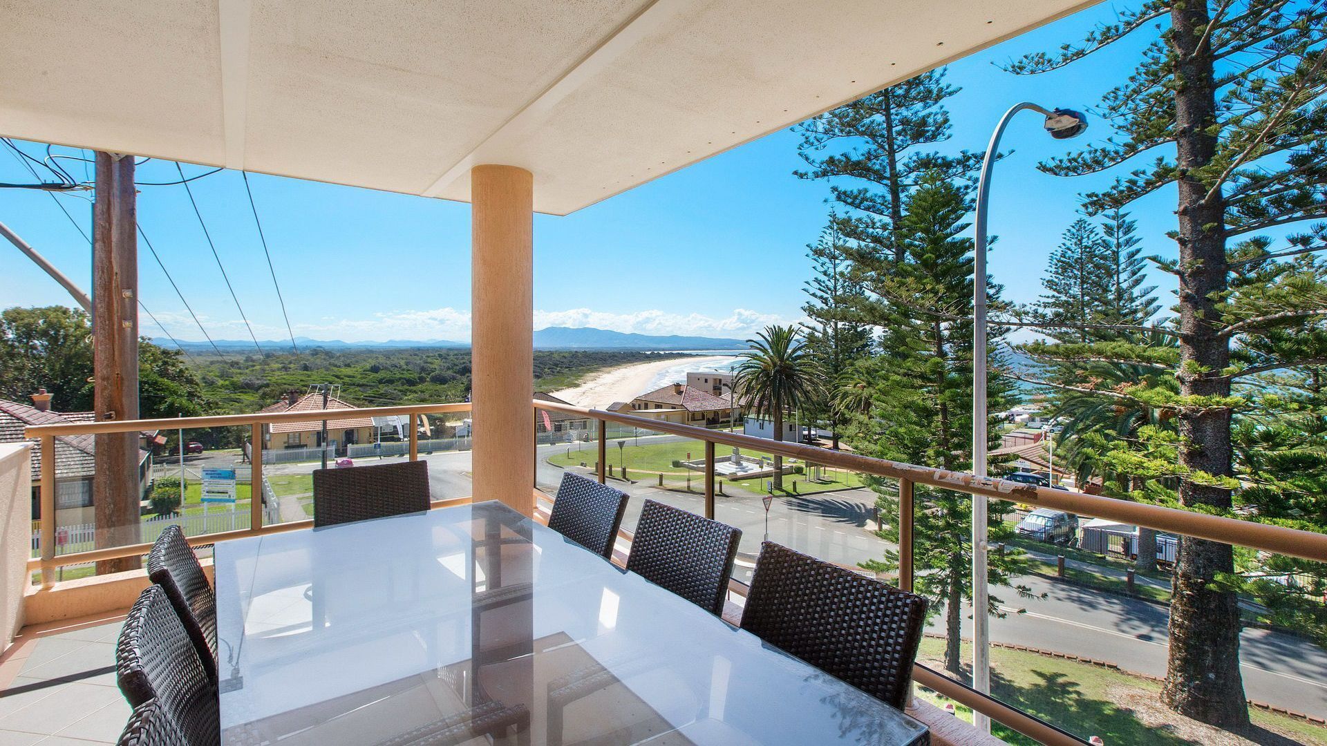 A balcony with a glass table and wicker chairs, overlooking a beach and mountains.