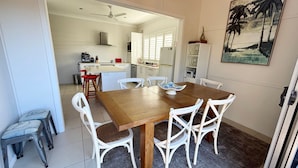 Un comedor con una mesa de madera y sillas blancas, una cocina al fondo y un cuadro en la pared.