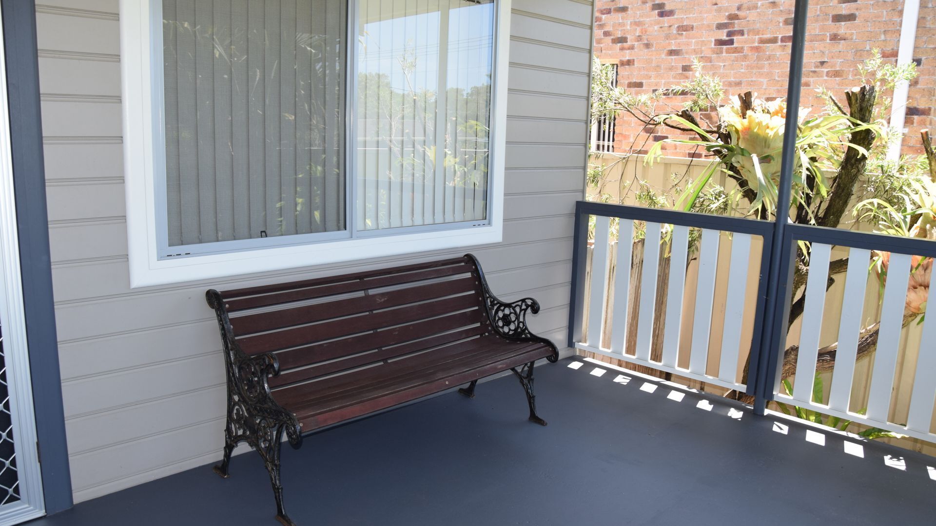 A wooden bench on a balcony with a view of a brick wall and a plant.