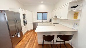 A modern kitchen with white cabinetry, a stainless steel refrigerator, a built-in oven, a sink, and a window with blinds. - Captains Cottage SWR (SOUTH WEST ROCKS)