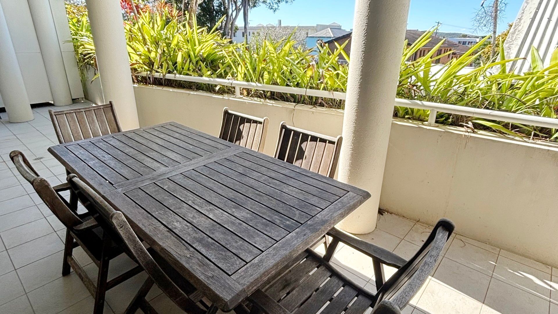 A wooden table with chairs on a balcony with a view of greenery and buildings.