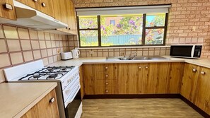 A kitchen with wooden cabinets, a gas stove, a microwave, and a window with a view of greenery. - Beach Shack SWR - Walk to the beach (SOUTH WEST ROCKS)