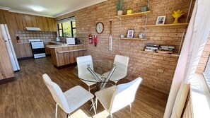 A modern kitchen with wooden cabinets, a built-in oven, and a dining area with a glass table and white chairs. - Beach Shack SWR - Walk to the beach (SOUTH WEST ROCKS)