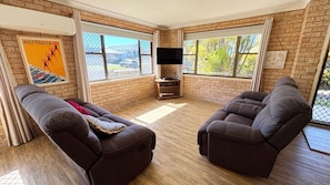A room with two reclining sofas, a flat-screen TV, and large windows with a view of the outside. - Beach Shack SWR - Walk to the beach (SOUTH WEST ROCKS)