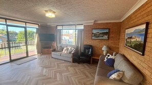 A spacious living room with a herringbone floor, a sofa, a chair, a TV, and a balcony with a view of houses. - Seacrest Unit 1 - walk to the beach (South West Rocks)