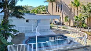A pool area with a white fence, a small building with a sign, and palm trees. - Ocean Shores Apartment 13 (SOUTH WEST ROCKS)